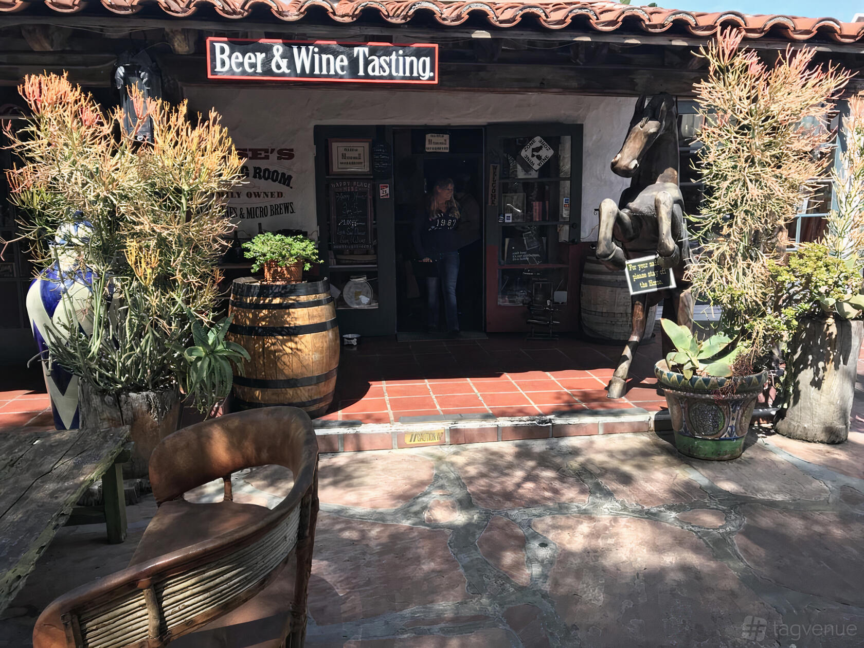 A rustic pub entrance with wooden barrels, potted plants, and a sculpture at Rose's Tasting Room.