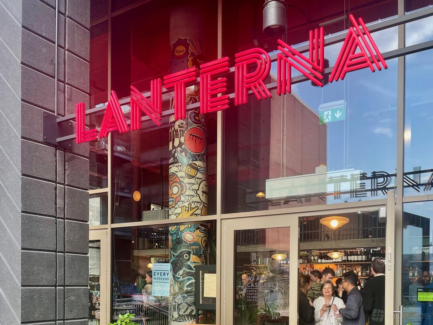 A bar with large glass windows, neon signage, and a mural-covered pillar at Lanterna Fish Island.