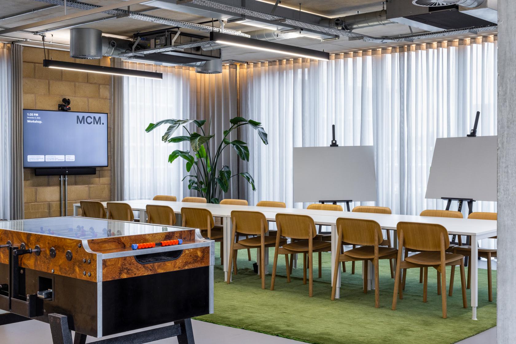 A coworking meeting room with long tables, wooden chairs, a foosball table, and large windows at The Studios. SE1.