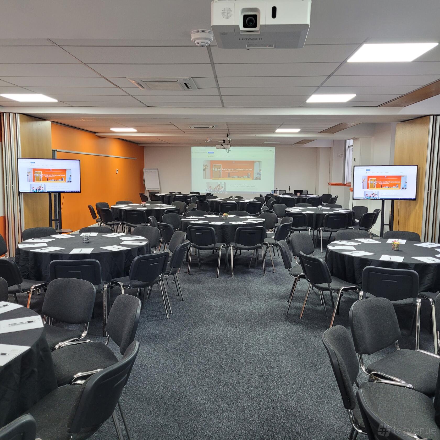 A meeting centre with round tables, black chairs, two projection screens, and flip charts at Edinburgh Training and Conference Venue.
