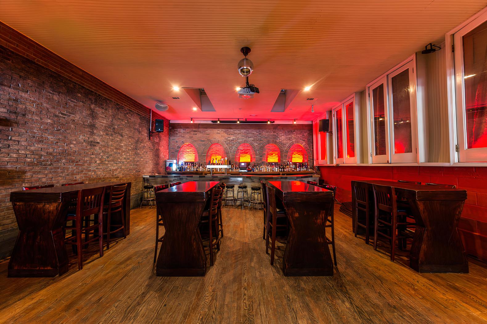 An event space with exposed brick walls, wood floors, dark tables, and a backlit bar at Johnny Pistolas.