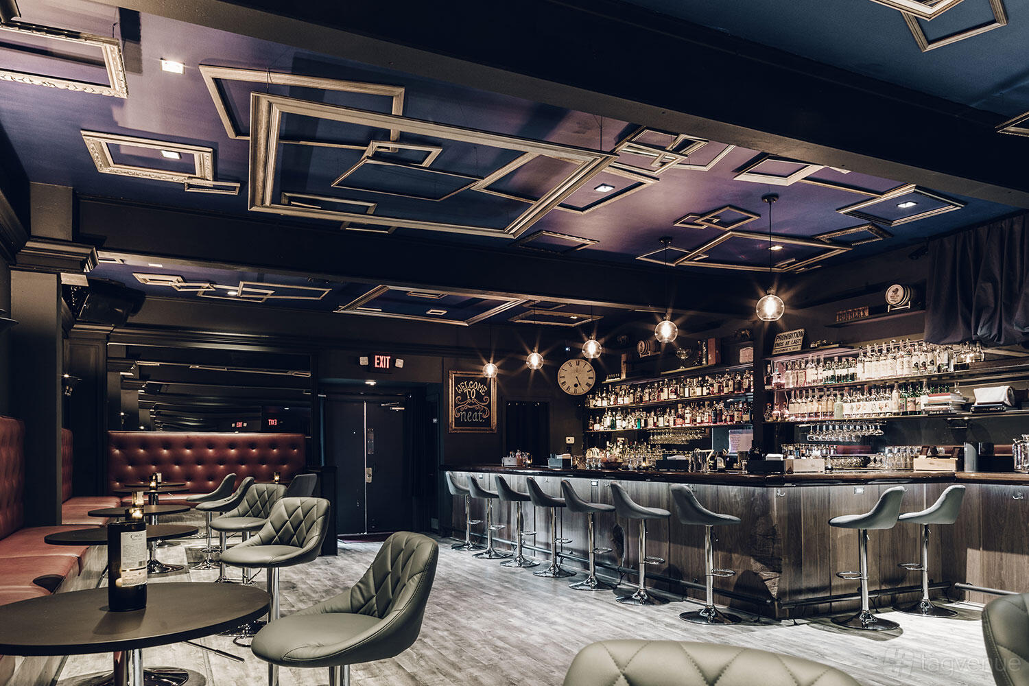 A cocktail bar with geometric ceiling accents, tufted banquette seating, and a sleek bar with high stools at neat.