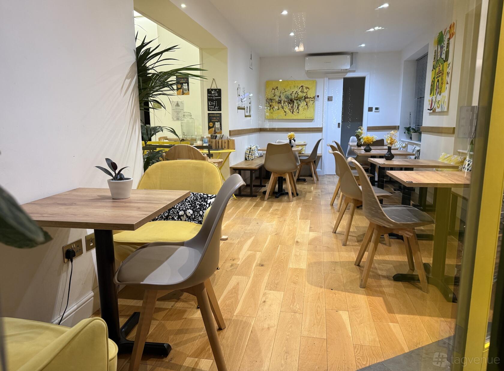 A cafe with wooden floors, yellow and gray chairs, potted plants, and wall art at Kofi Cafe.