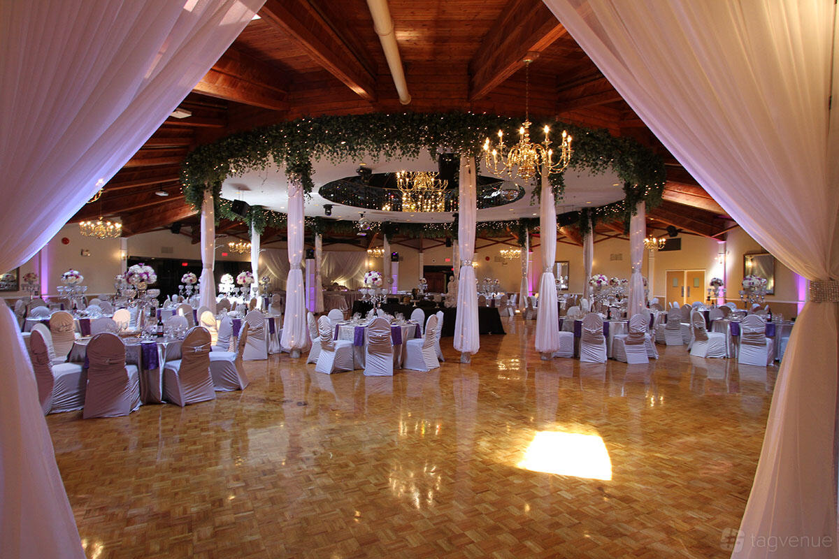 A banquet hall with draped entry, round tables with white linens, chandeliers, and greenery ceiling accents at Oakville Legacy Banquet & Convention Centre.