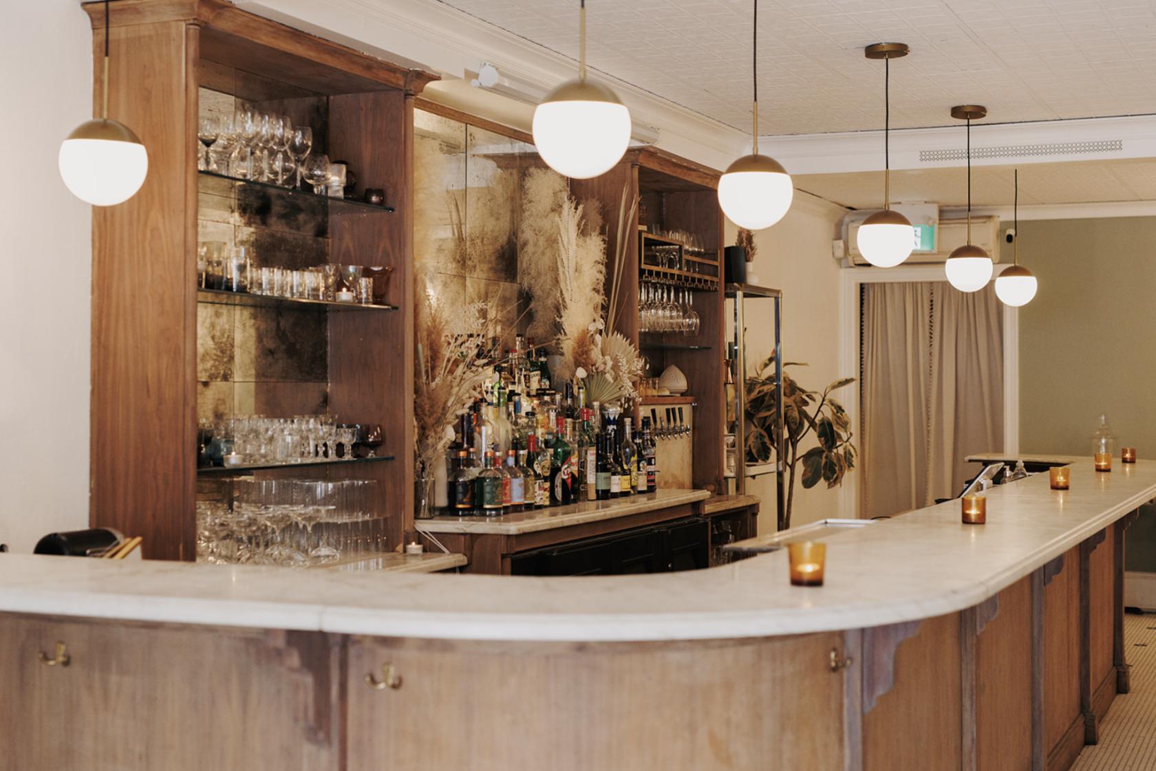 An event venue with a marble-topped wooden bar, glassware shelves, globe pendant lights, and dried floral decor at Ahma Toronto.