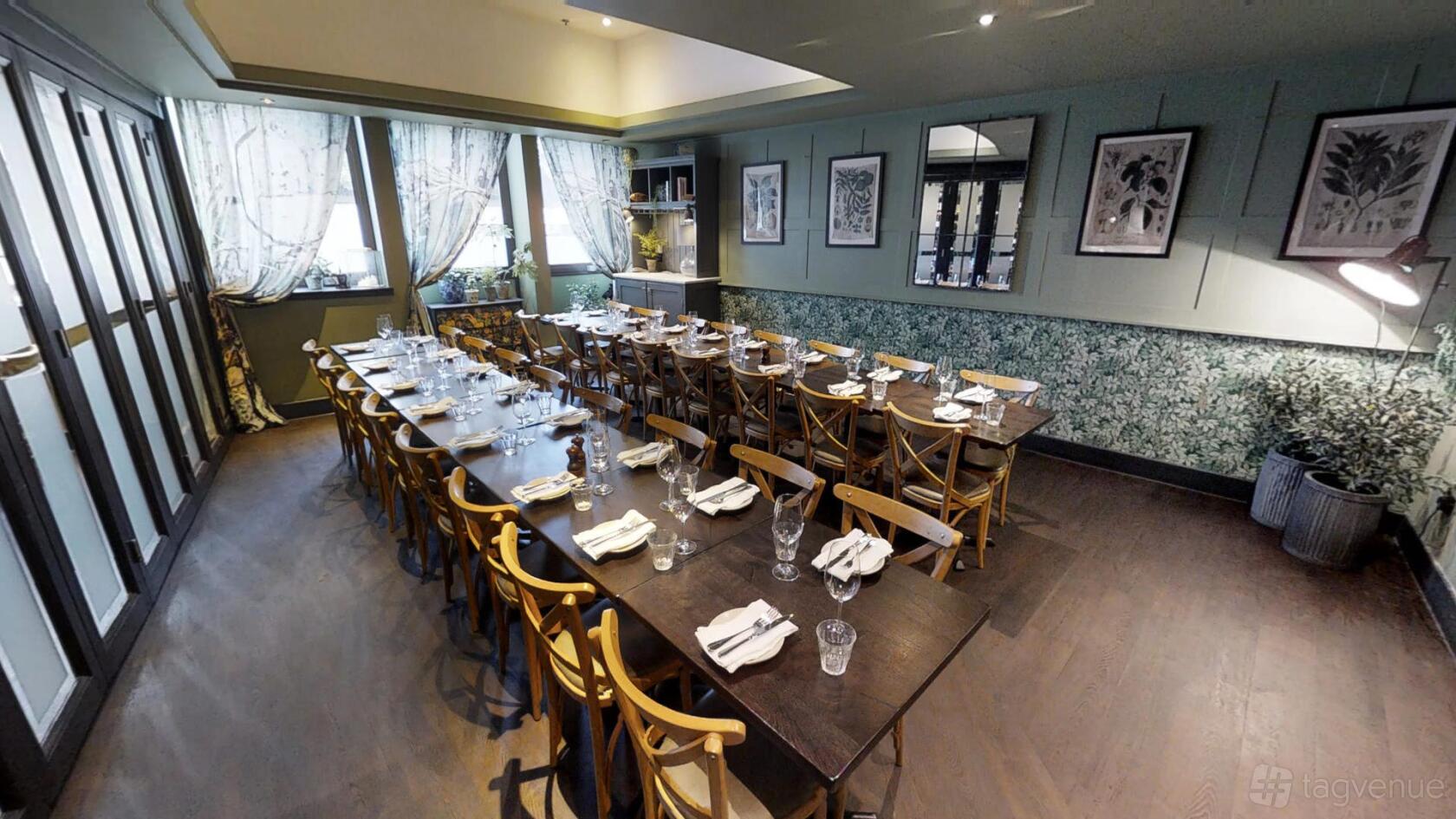 A dining room with long wooden tables, cross-back chairs, framed art, and floral wallpaper at Brasserie Blanc Southbank.
