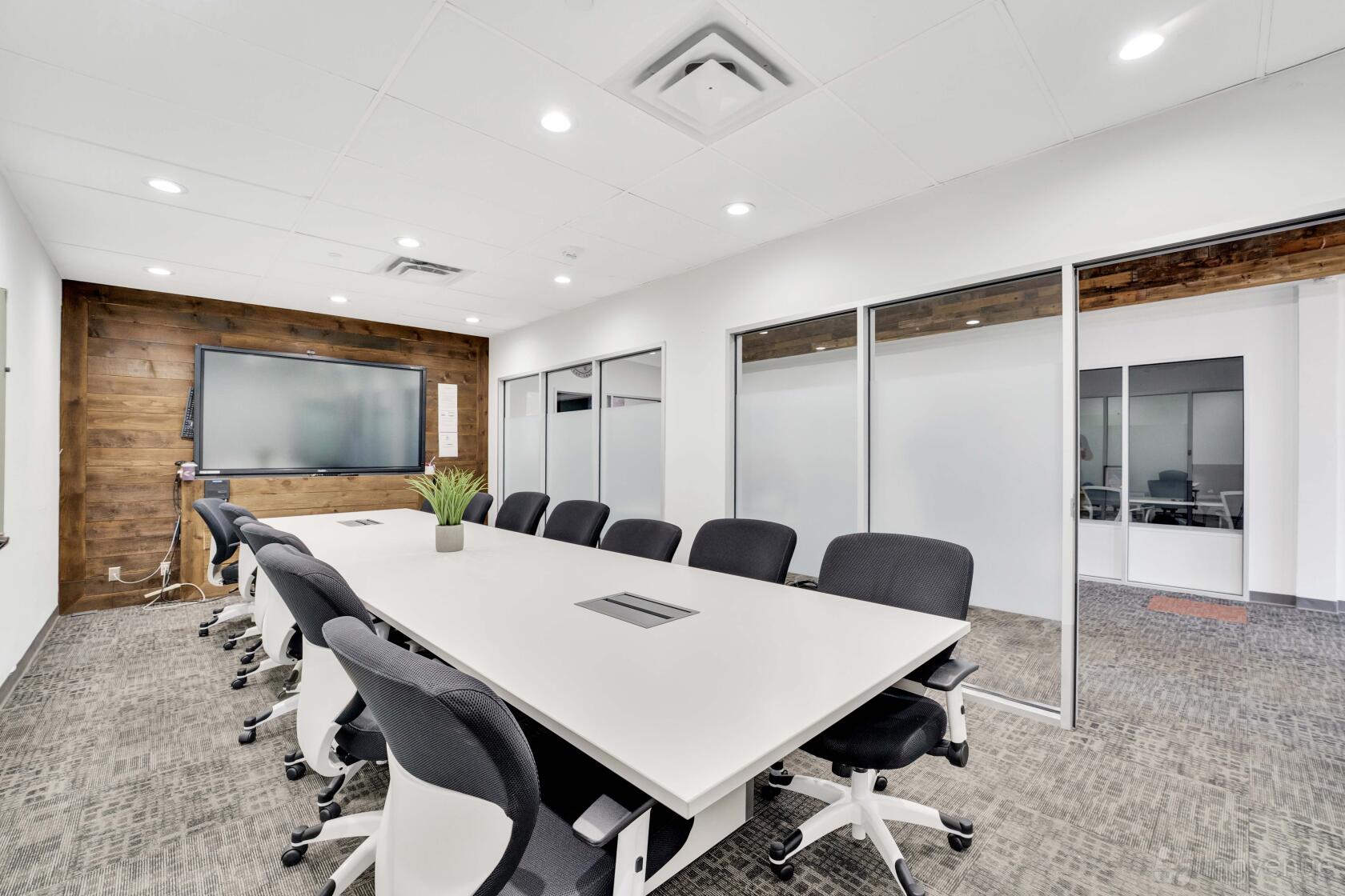 A coworking space conference room with a large white table, black chairs, wood accent wall, and wall-mounted screen at The Drawing Board.