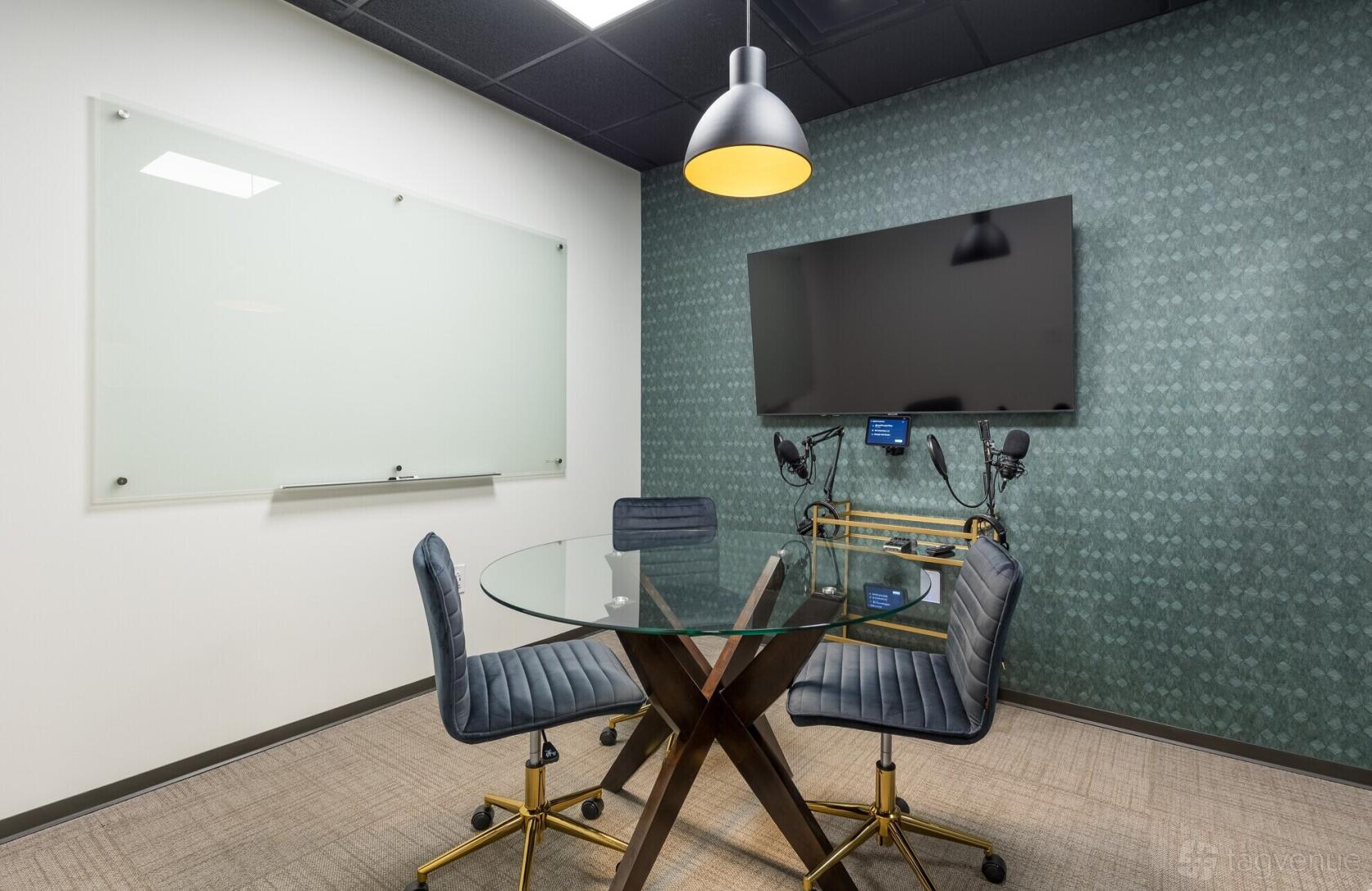 A small meeting room with a round glass table, three blue chairs, wall-mounted screen, and whiteboard at Lucid Private Offices - Cumberland.