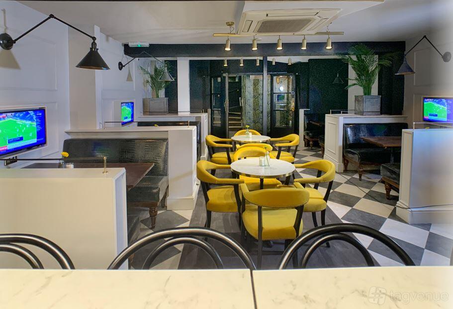 A lounge in a pub with yellow upholstered chairs, marble-topped tables, chequered flooring, and wall-mounted TVs at Bonds.