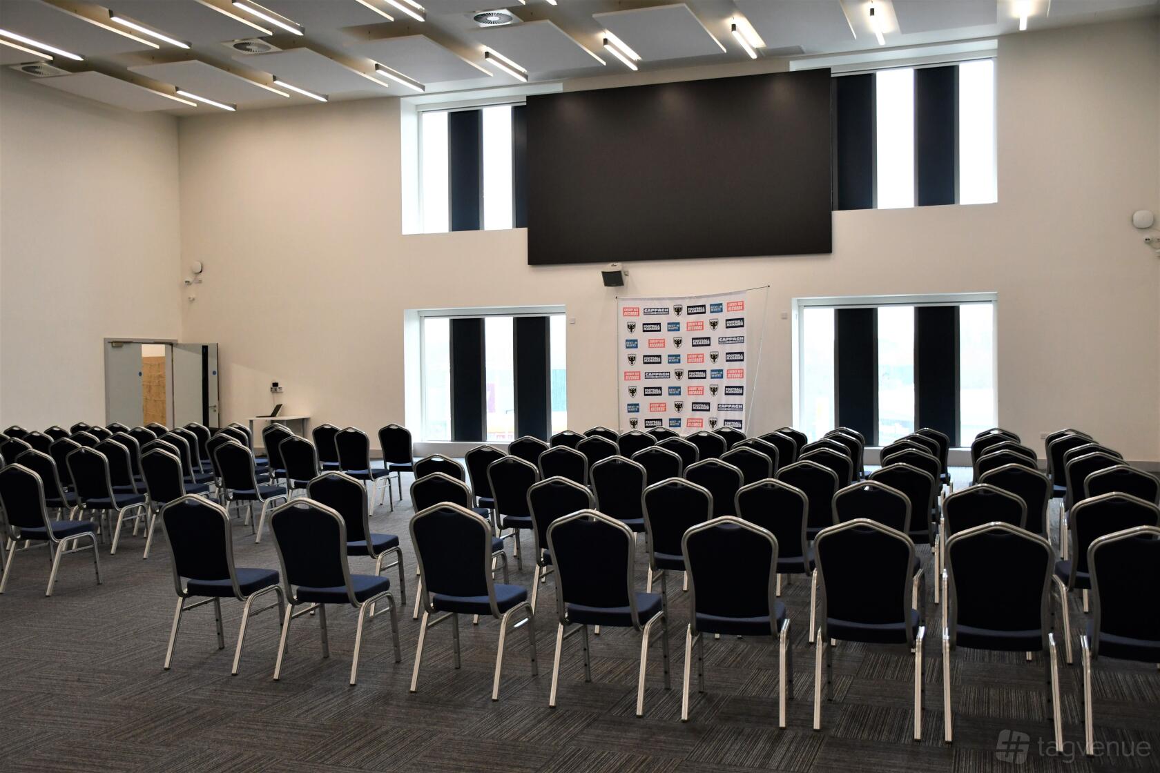 An event space with rows of black chairs, a projector screen, and tall windows at AFC Wimbledon Plough Lane Stadium C&E.