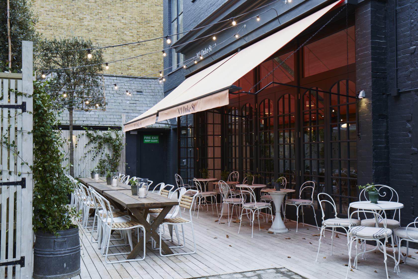 An outdoor pub courtyard with string lights, wooden decking, long tables, and metal chairs at No.1 Duke Street.