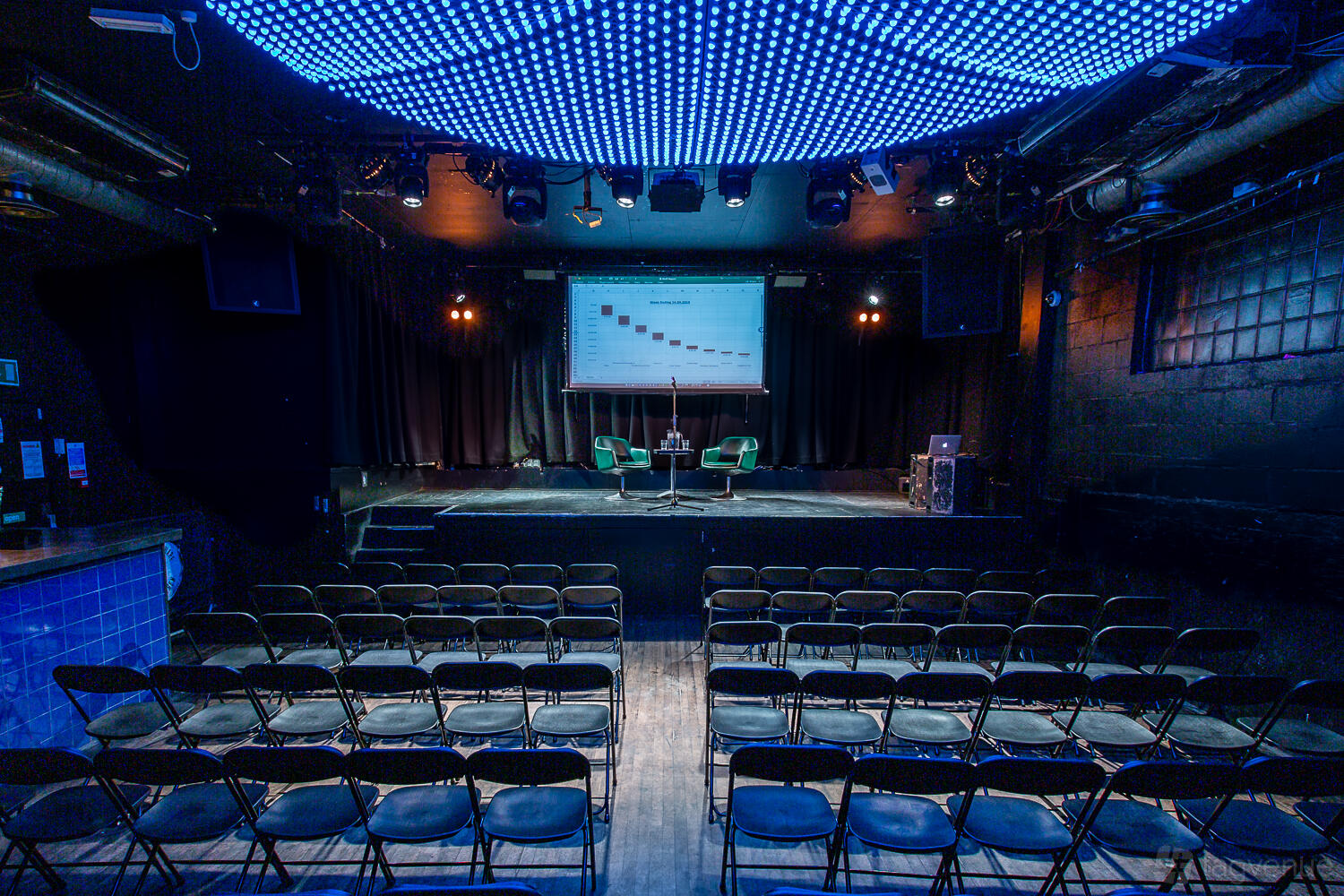 An event space with a stage, rows of chairs, black walls, and LED ceiling lights at Colours Hoxton.