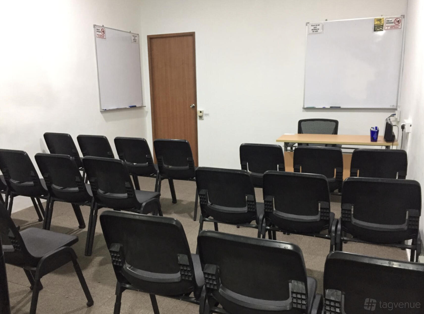 A training room with rows of black chairs facing a desk, two whiteboards, and a wooden door at Synergy Room Singapore Shopping Centre.