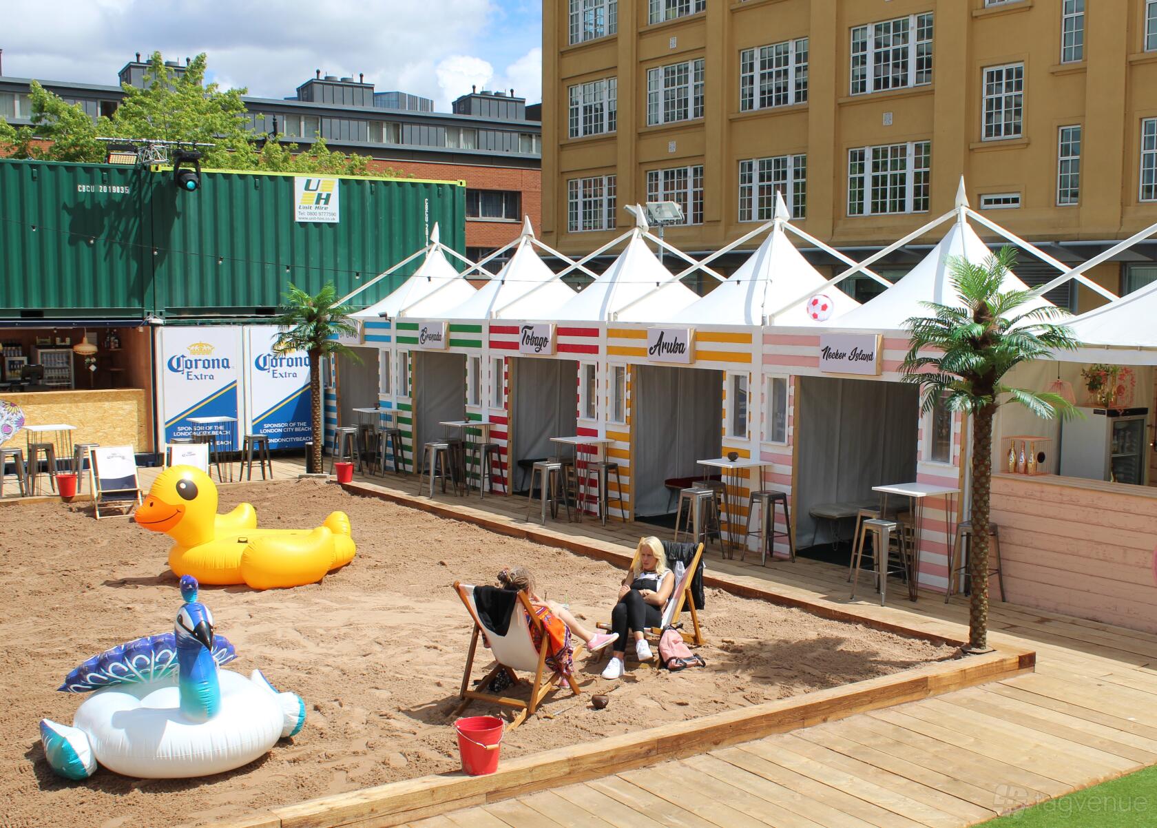 An outdoor event venue with a sandy area, inflatable pool floats, white cabanas, and deck chairs at The London City Beach.