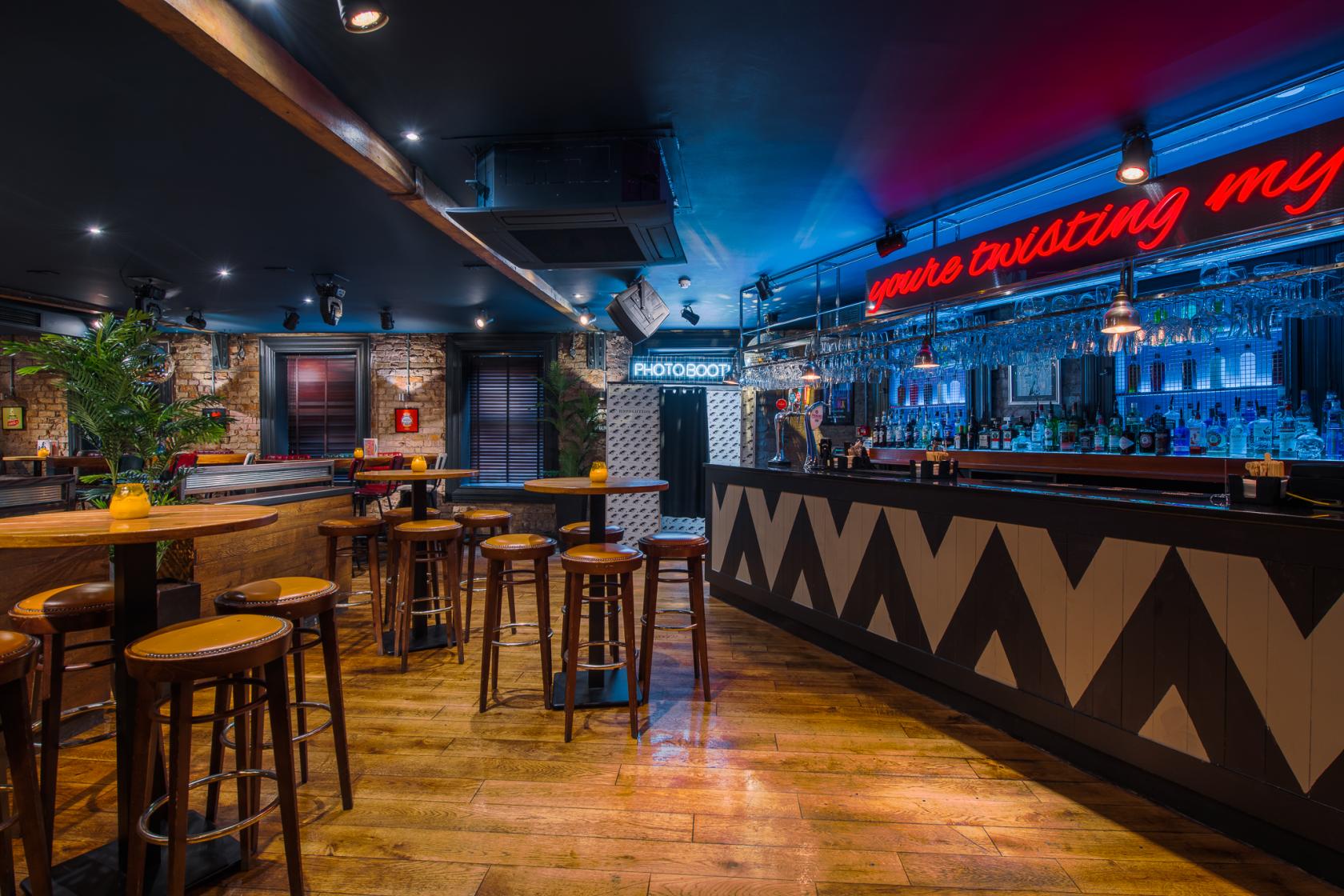 A bar with exposed brick walls, neon signage, wooden flooring, and high tables with stools at Revolution Oxford Road.
