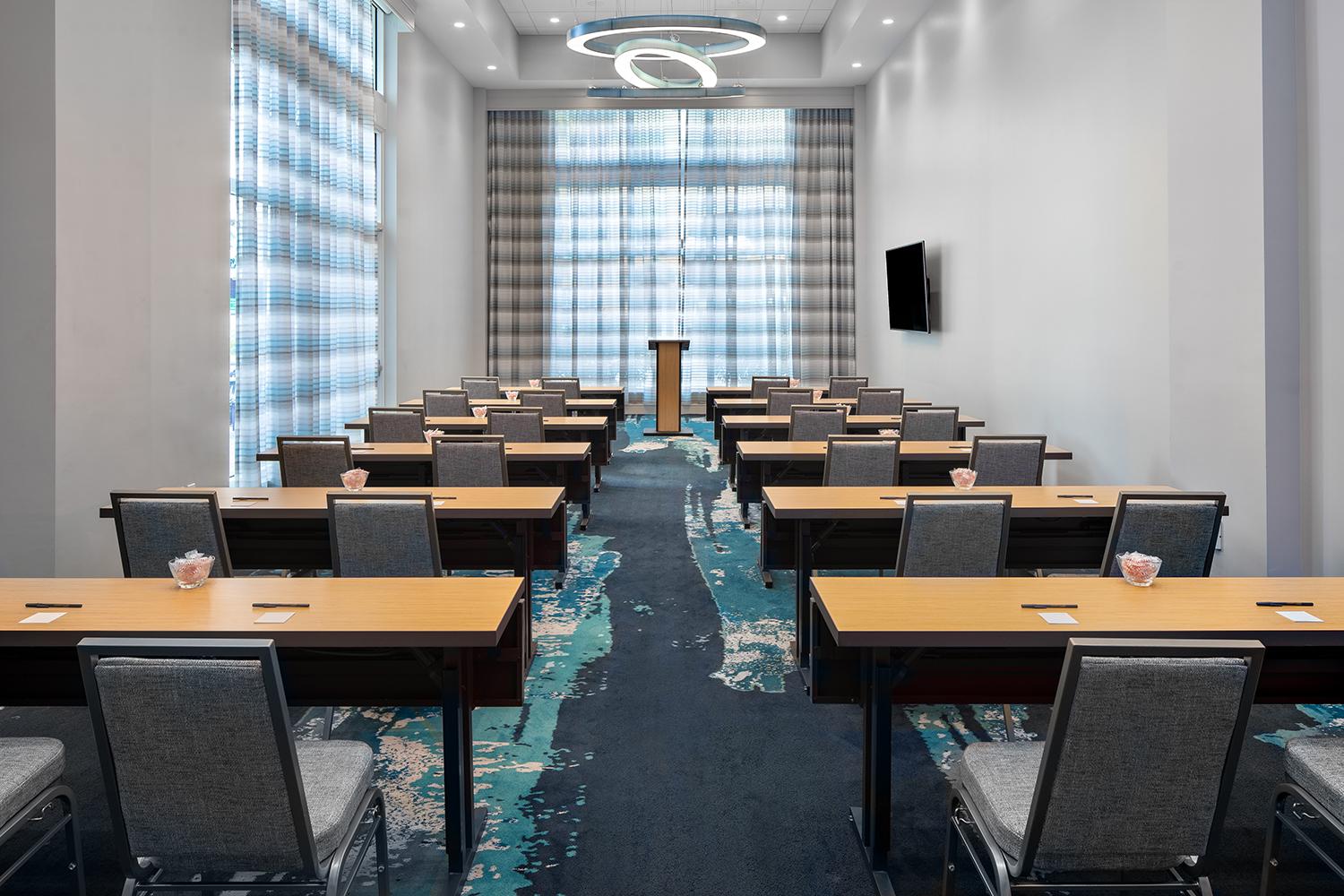 A meeting room with classroom-style tables, upholstered chairs, modern lighting, and floor-to-ceiling windows at Courtyard Delray Beach.