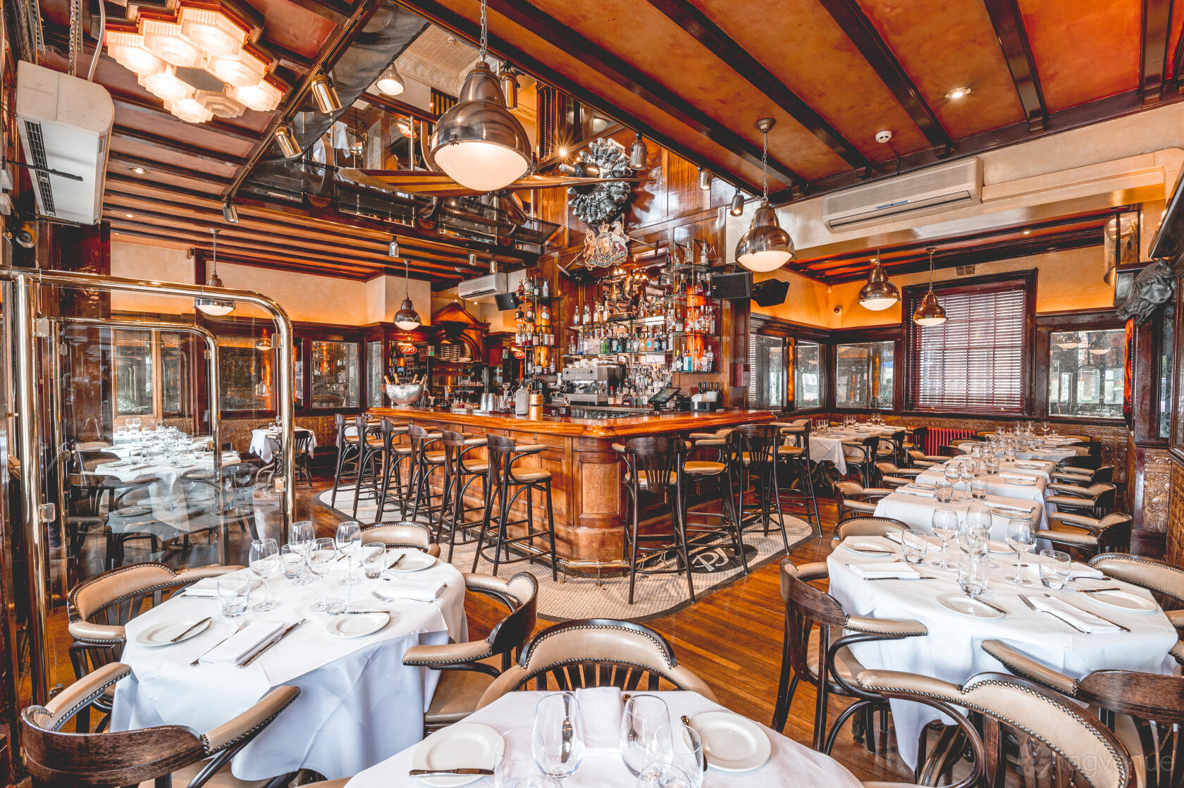 A restaurant with white-linen round tables, wood-paneled walls, and a central curved bar at PJ's Chelsea Brasserie.
