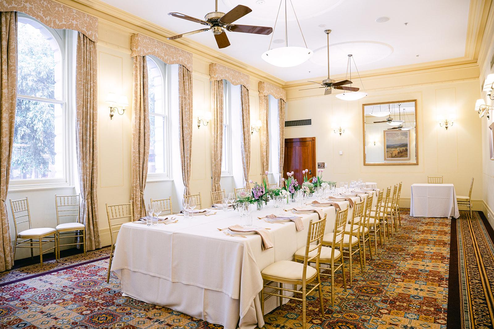 A function room with large windows, ornate curtains, chandeliers, and a long banquet table at The Hotel Windsor.