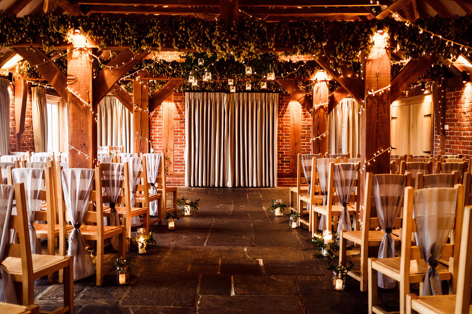A barn venue with exposed timber beams, draped curtains, and ceremony seating at The Ferry House.