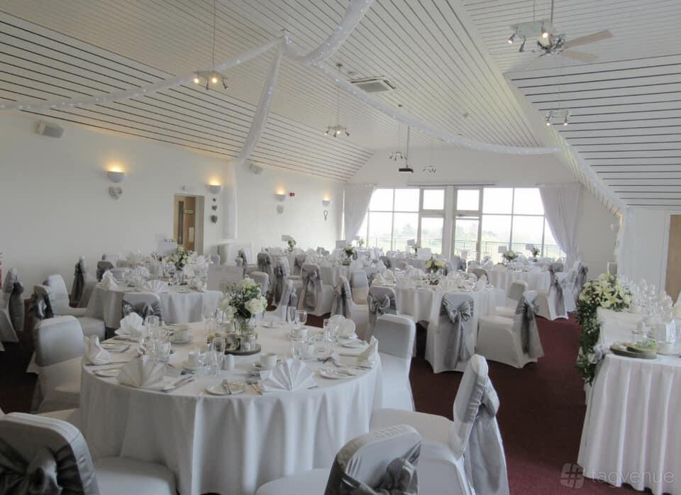 An event space with round tables draped in white linens, chair covers with bows, and large windows at Ufford Park Woodbridge.