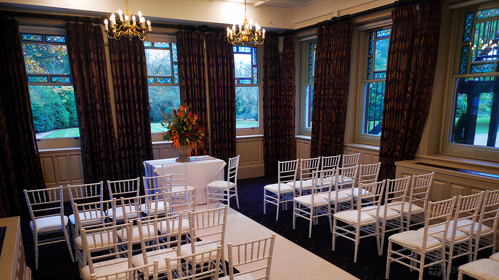An event room with white chairs, chandeliers, tall windows, and dark patterned curtains at Woodlands Park Hotel
