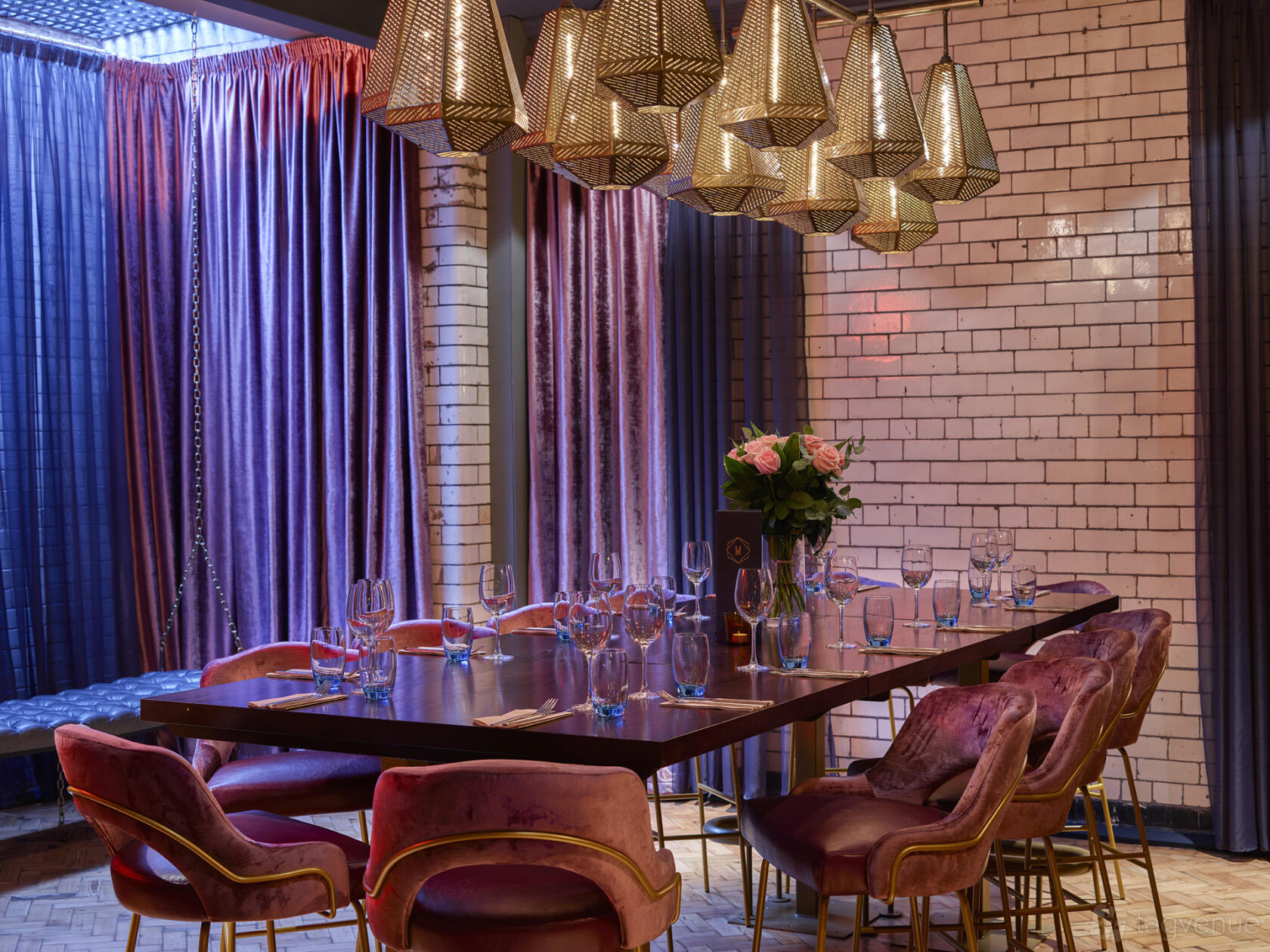 A dining room with velvet chairs, a long wooden table, gold pendant lights, and white tiled walls at Manahatta Deansgate.