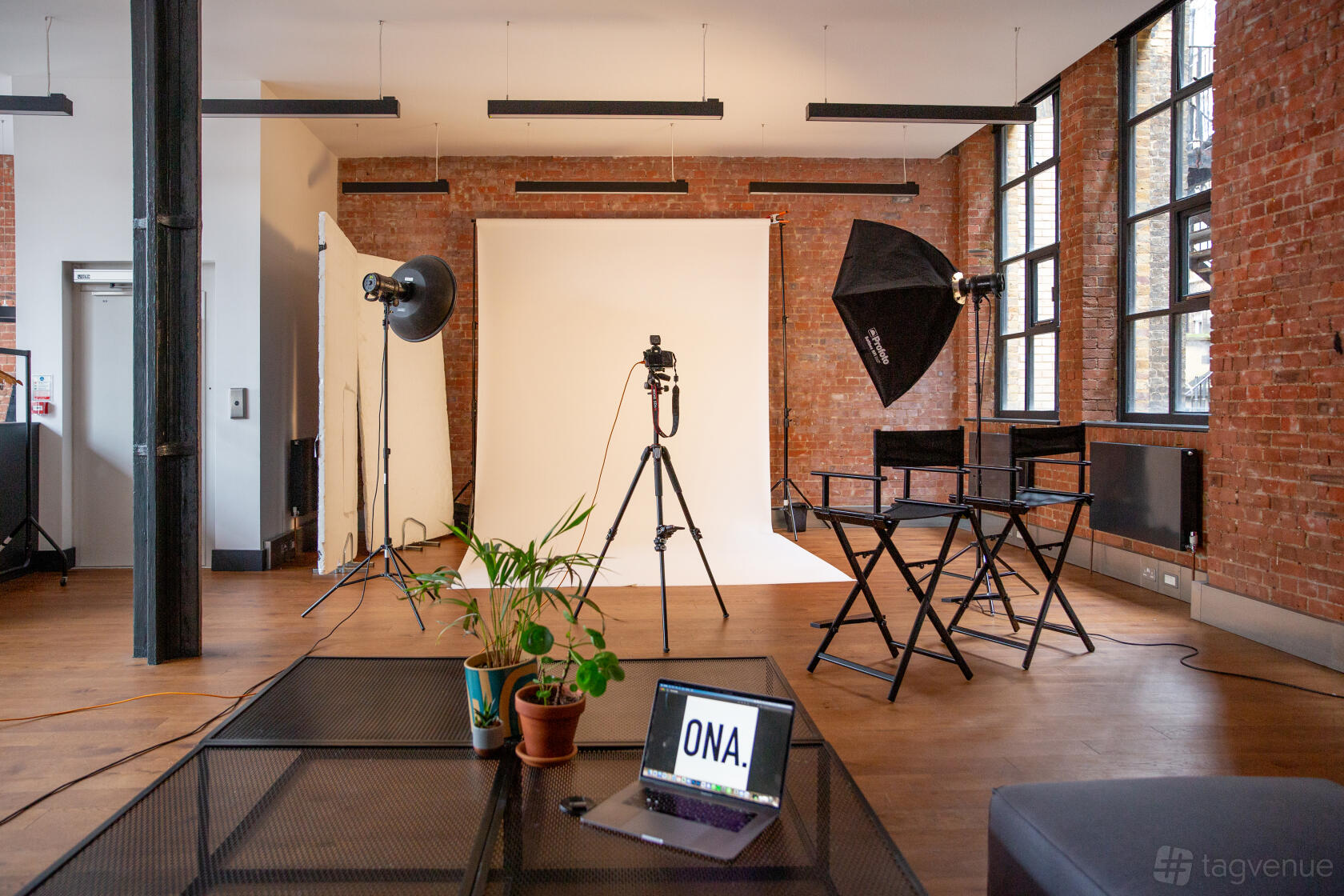 A photo studio with exposed brick walls, large windows, studio lighting equipment, and a backdrop at Ona. Studio - Photo / Video Studio.