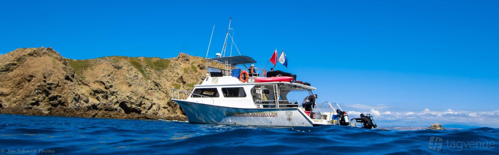 A charter boat with open decks, safety equipment, and flags moored near rocky shore at Waterhorse Charters.