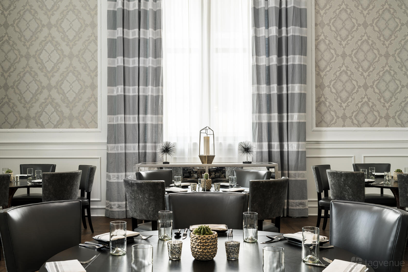 A dining room with round black tables, leather chairs, and tall windows with patterned drapes at Aqimero.