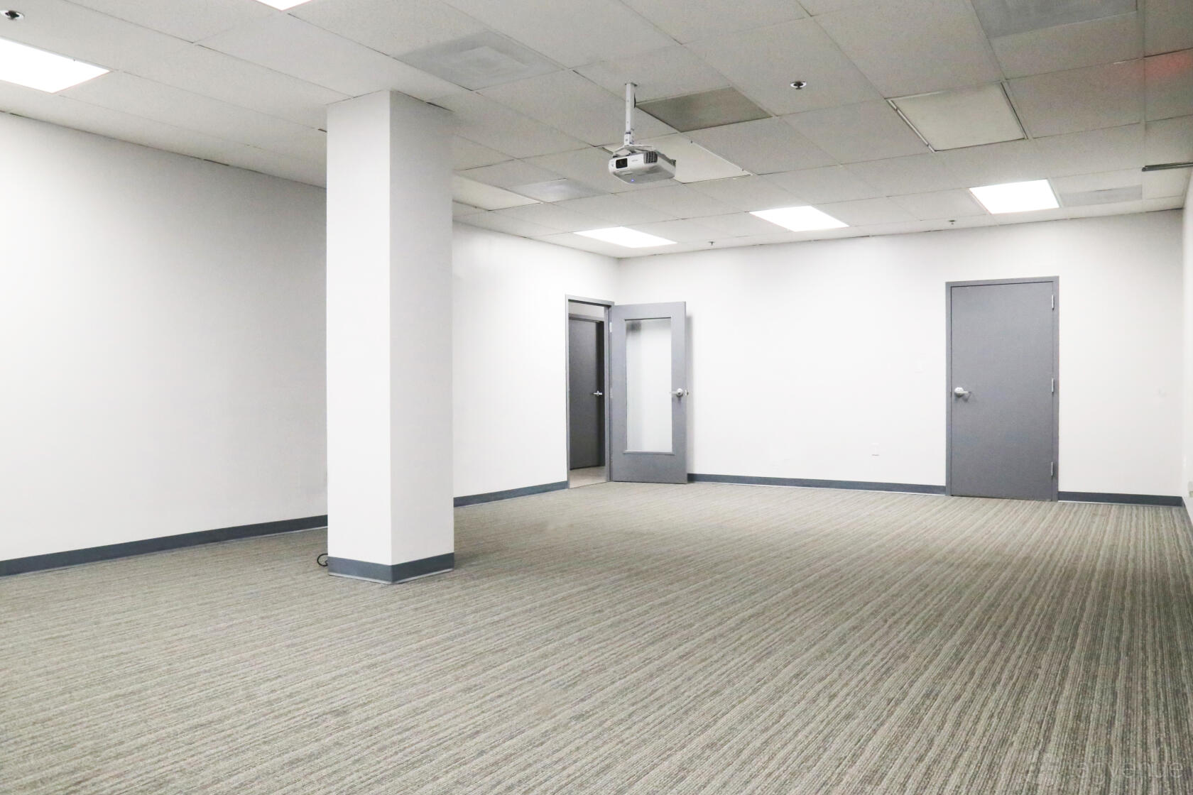 An event space in a coworking office with neutral walls, carpeted floor, and ceiling-mounted projector at Perfect Office Solutions - Riverdale 6811.