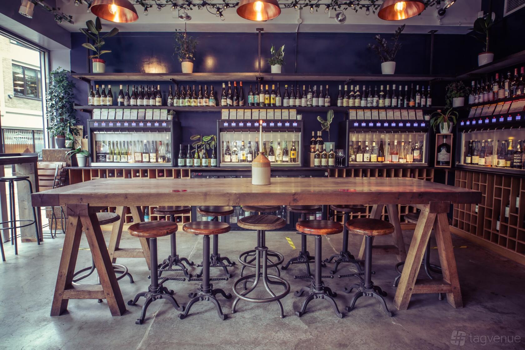 A bar with a long wooden communal table, industrial stools, hanging lights, and wine bottle displays at Vagabond Fulham.