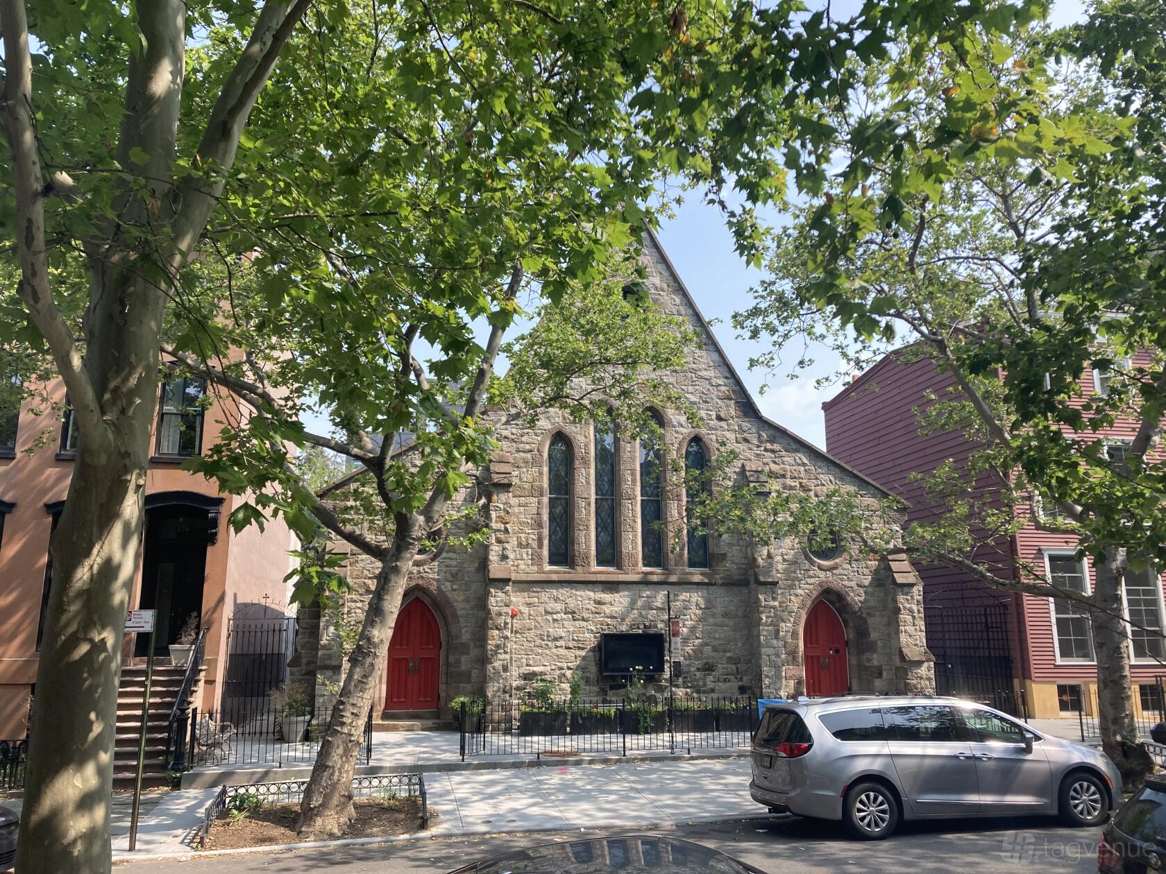 A historic stone church with tall arched windows, red doors, and leafy trees at Church of the Ascension.