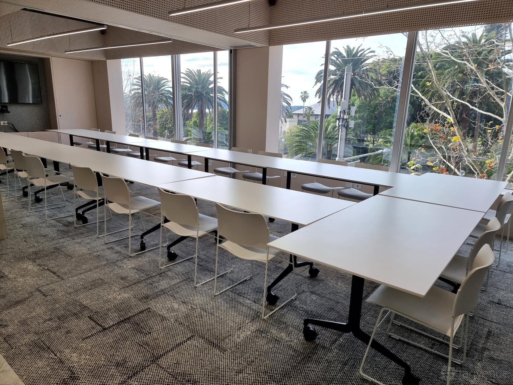 A meeting centre with rows of white tables, white chairs, and floor-to-ceiling windows overlooking palm trees at FlexSpot Macquarie.