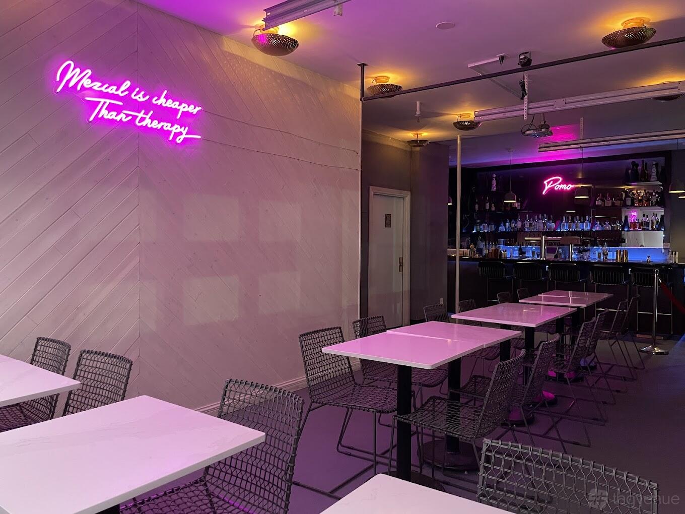 A restaurant with pink neon signage, wire chairs, white tables, and a backlit bar at Pomo Brooklyn.