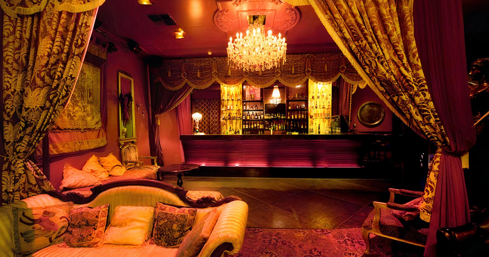 A nightclub dining room with velvet drapes, ornate chandeliers, plush seating, and a backlit bar at Chasers Nightclub and Venues.