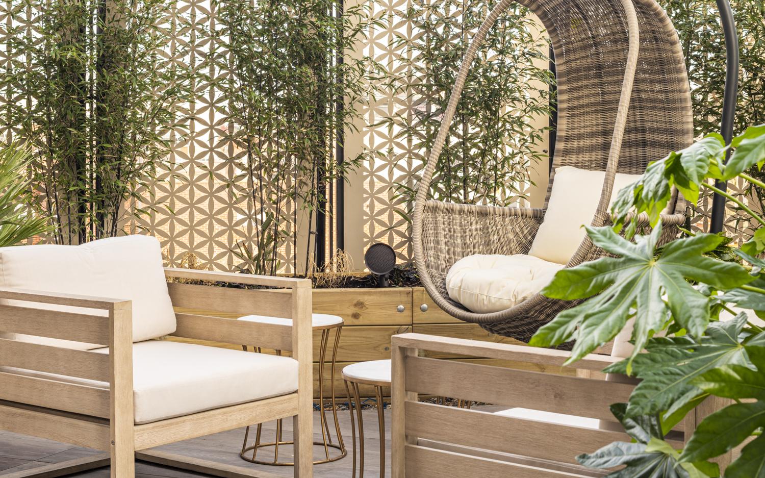 A hotel terrace with wooden lounge seating, round side tables, a hanging wicker chair, and potted plants at The Gate London City.