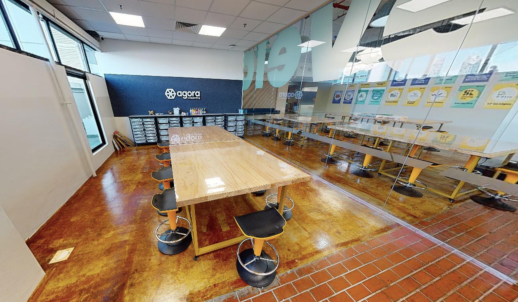 A meeting room with long wooden tables, stools, and storage bins beneath a glass wall at Agora Colearning.