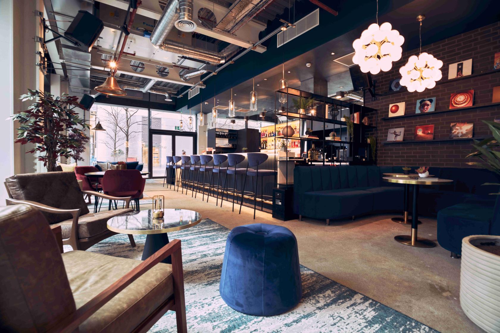 A cocktail bar lounge with velvet seating, large windows, vinyl records on the wall, and a marble-topped bar at Qube East - Cocktail Bar & Recording Studios.