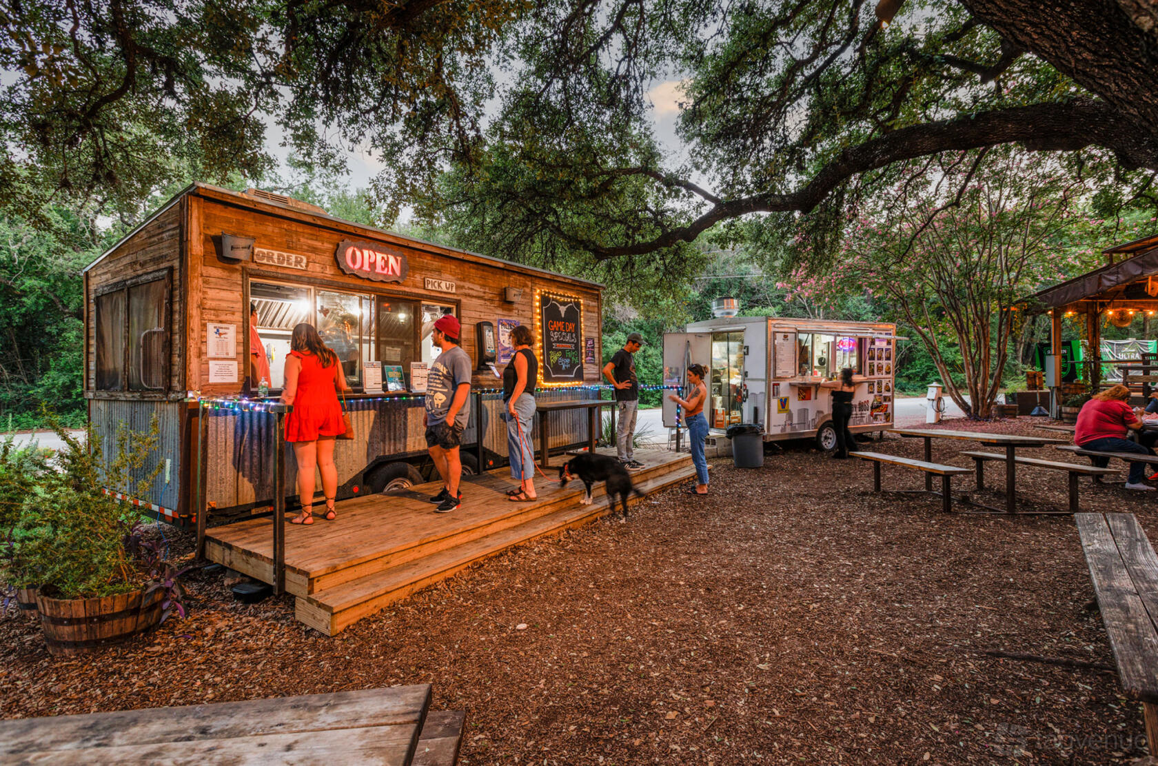 An outdoor bar area with picnic tables, food trucks, string lights, and large shade trees at Moontower Saloon.