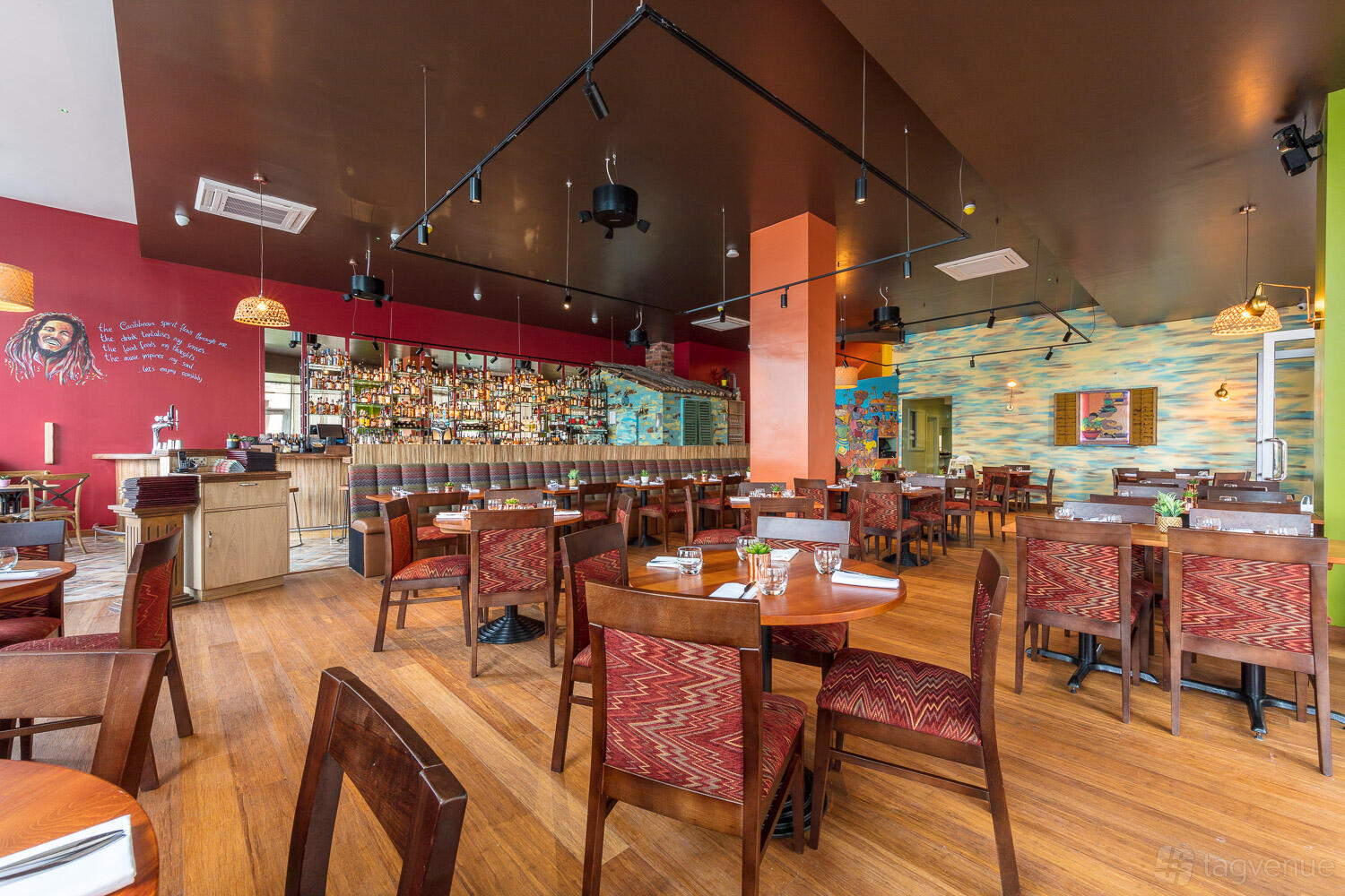 A restaurant with wooden floors, round tables, colorful wall art, and a stocked backlit bar at Cottons Vauxhall.