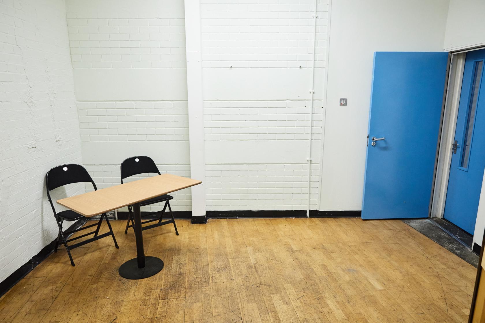 A rehearsal studio with wood flooring, white painted brick walls, two folding chairs, and a small table at Seven Dials Playhouse.