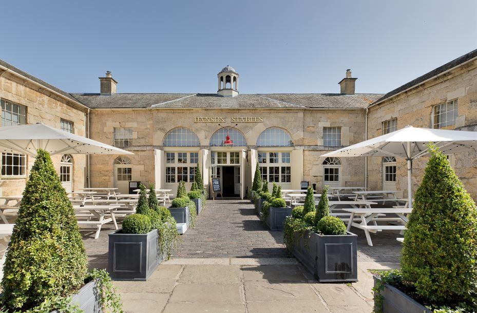 A pub courtyard with wooden picnic tables, large white umbrellas, and manicured hedge planters at Danson Stables.