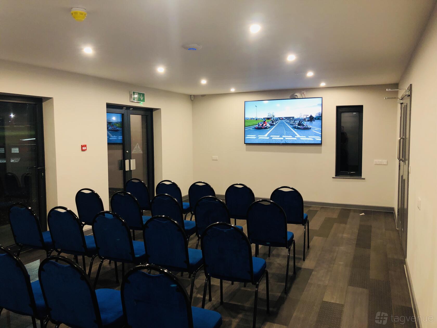 An event space with rows of blue chairs facing a wall-mounted screen and recessed lighting at Whilton Mill.