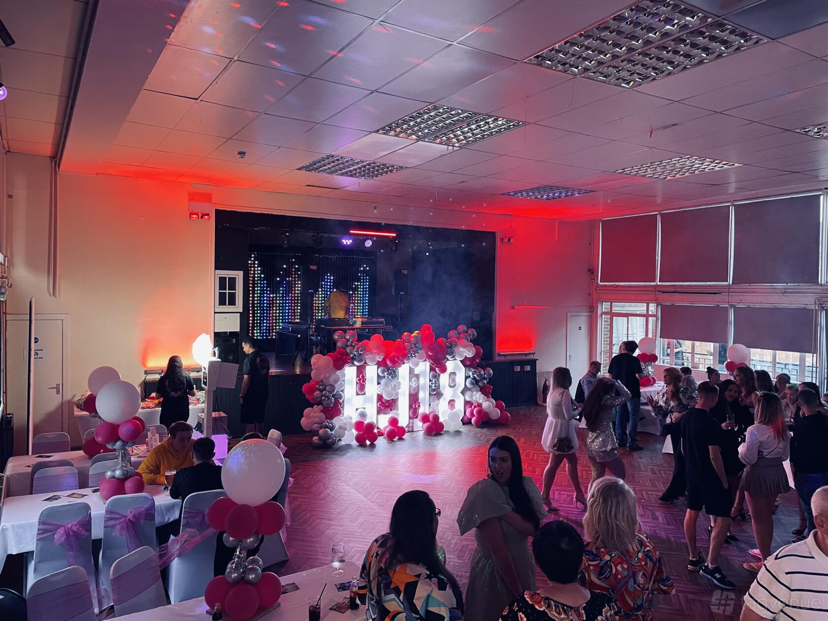 A hall with a decorated stage, pink and white balloons, and guests seated at tables at Magdalen Hall.