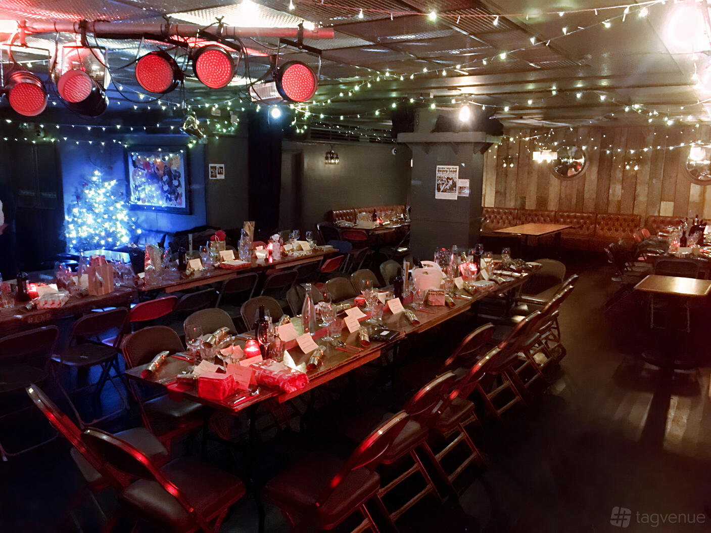 An underground pub space with long banquet tables, folding chairs, string lights, and a decorated Christmas tree at The Phoenix, Cavendish Square.