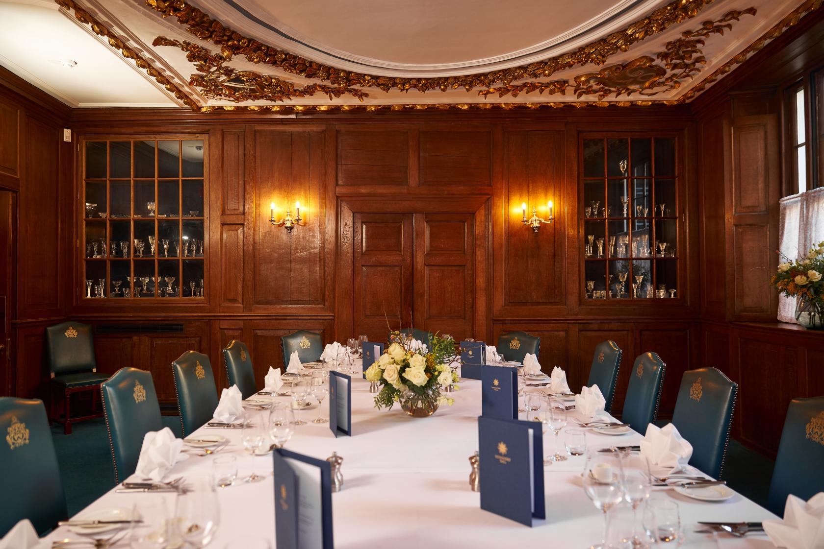 A dining room with wood panelled walls, glass-fronted cabinets, and a long table set for a meal at Innholders' Hall.