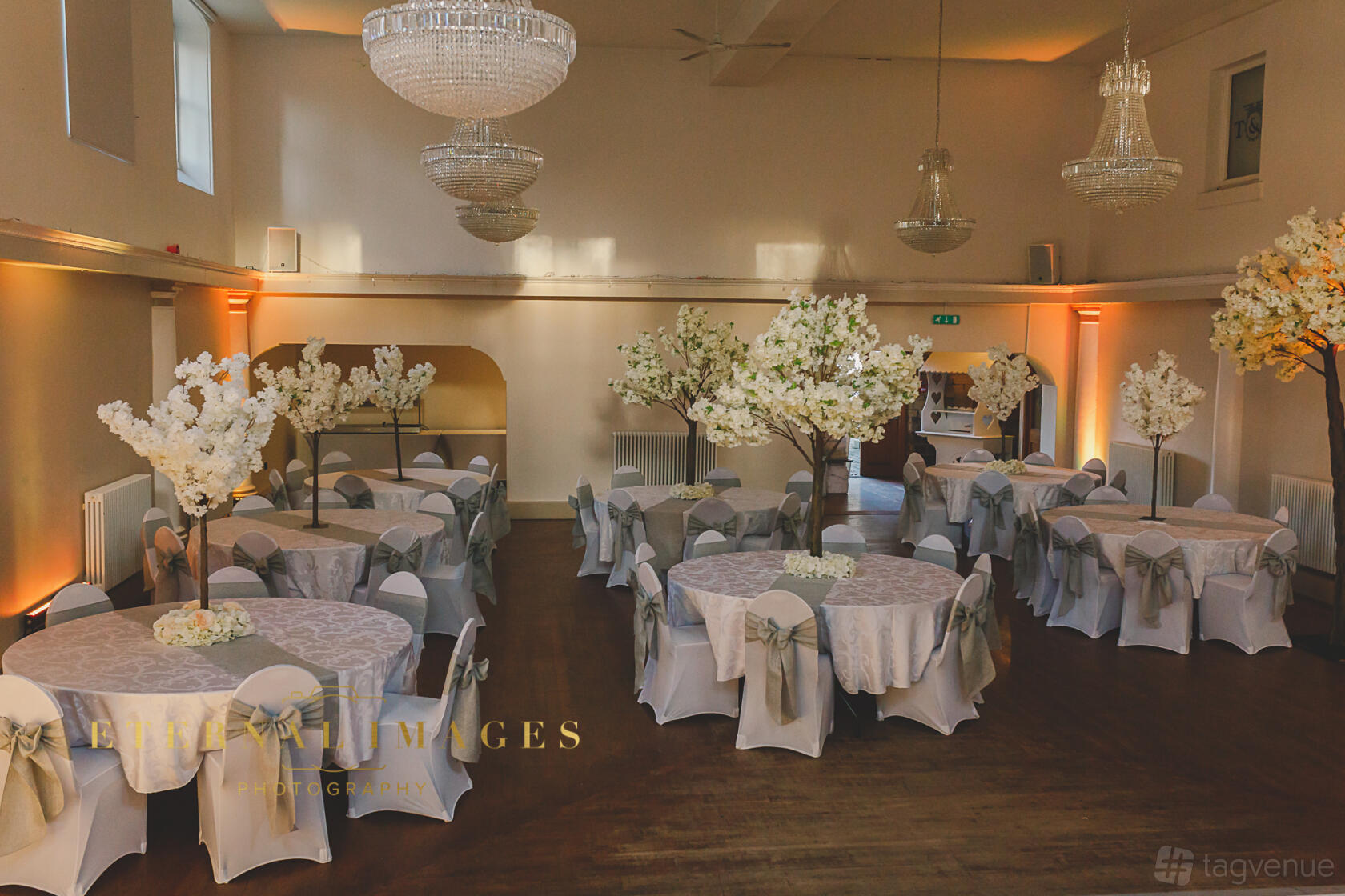 A ballroom hall with round tables draped in white linens, tall floral centerpieces, and crystal chandeliers at Wortley Hall.