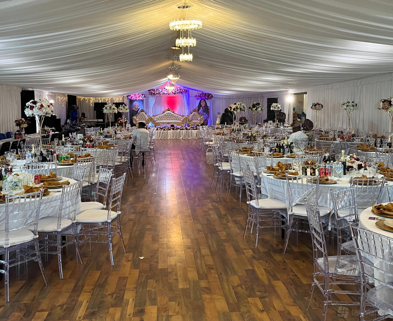 An event venue with draped white ceilings, chandeliers, clear chairs, and banquet tables at D’Will Banqueting Suite.
