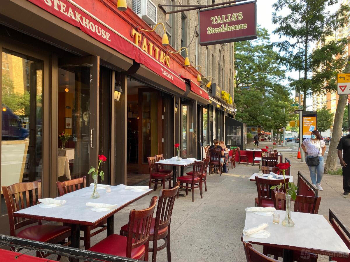 An outdoor restaurant seating area with red chairs, white tablecloths, and single roses at Talia's Steakhouse & Bar.