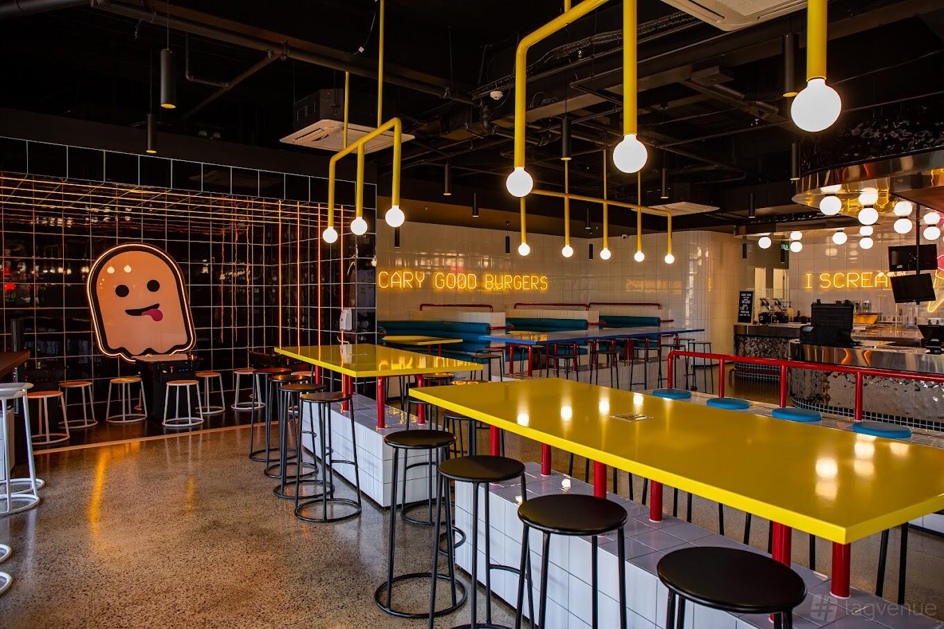 A restaurant with yellow communal tables, neon signage, and a cartoon ghost mural at Ghost Burgers and Grill.